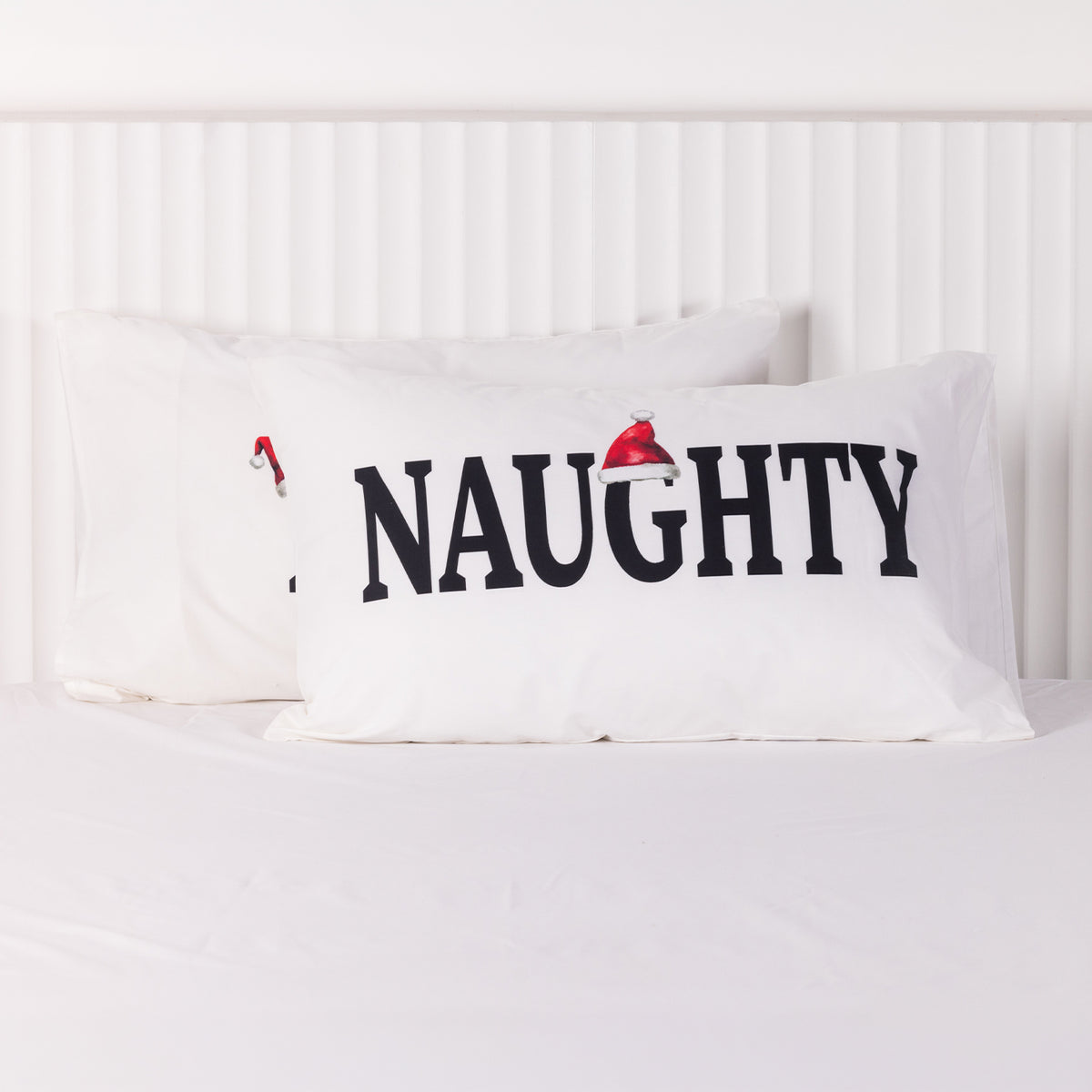 White Christmas Wishes Naughty and Nice pillowcases featuring bold black lettering and festive Santa hats, displayed overlapping on a white bed.