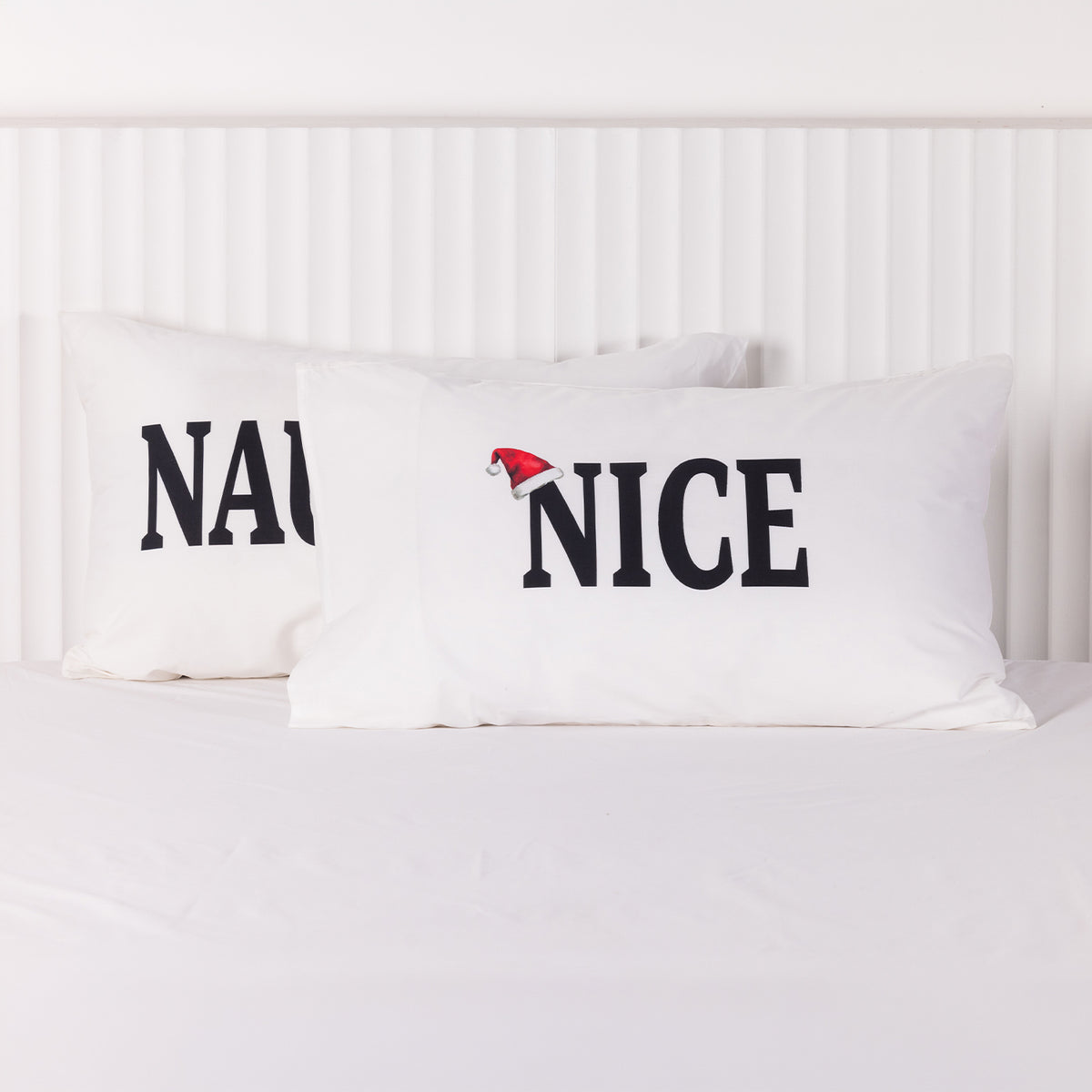 Close-up of the white Christmas Wishes Nice pillowcase featuring bold black lettering and a red Santa hat, displayed in front of the Naughty pillowcase.