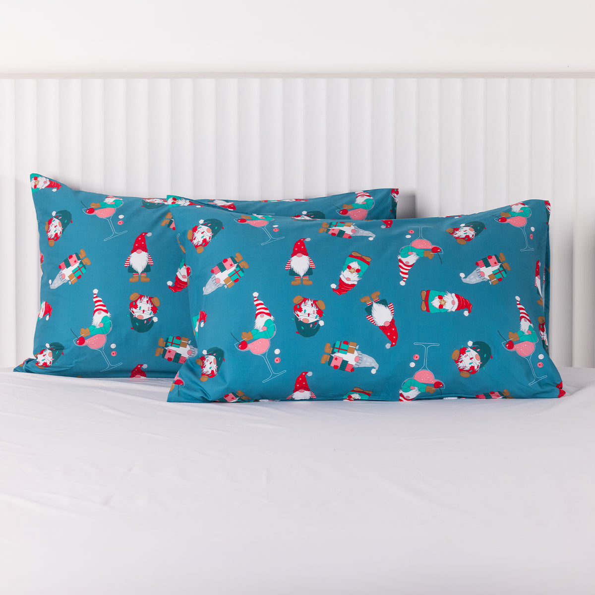 Two teal blue pillowcases featuring a playful Christmas gnome print with festive drinks and presents, displayed on a bed with white bedding.