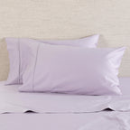 A Gracious Living Avery 750TC 100% Cotton Standard Pillowcase Pair in Dusky Mauve, styled on a bed with coordinating lavender-purple bedding. The pillowcases feature a smooth, elegant finish with subtle decorative stitched border detailing.
