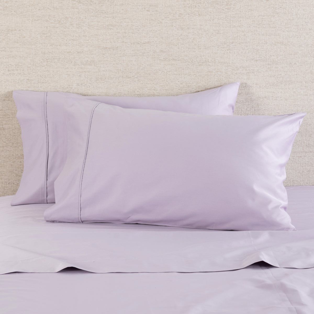 A Gracious Living Avery 750TC 100% Cotton Standard Pillowcase Pair in Dusky Mauve, styled on a bed with coordinating lavender-purple bedding. The pillowcases feature a smooth, elegant finish with subtle decorative stitched border detailing.