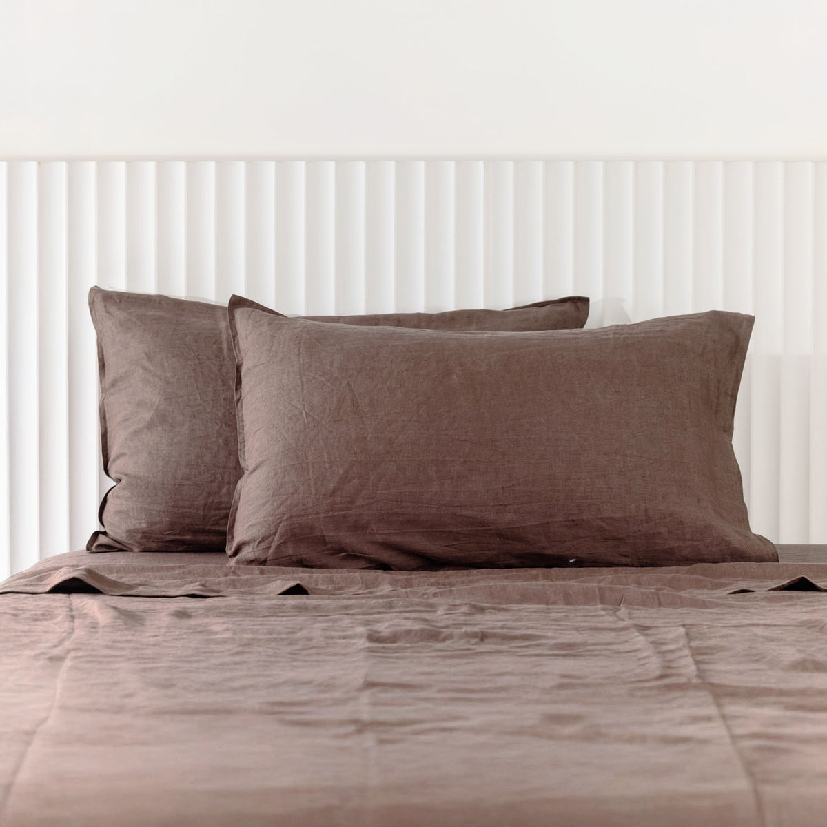 Cocoa linen pillowcases are beautifully layered against a white panelled headboard, showcasing their natural woven texture and relaxed, contemporary elegance.
