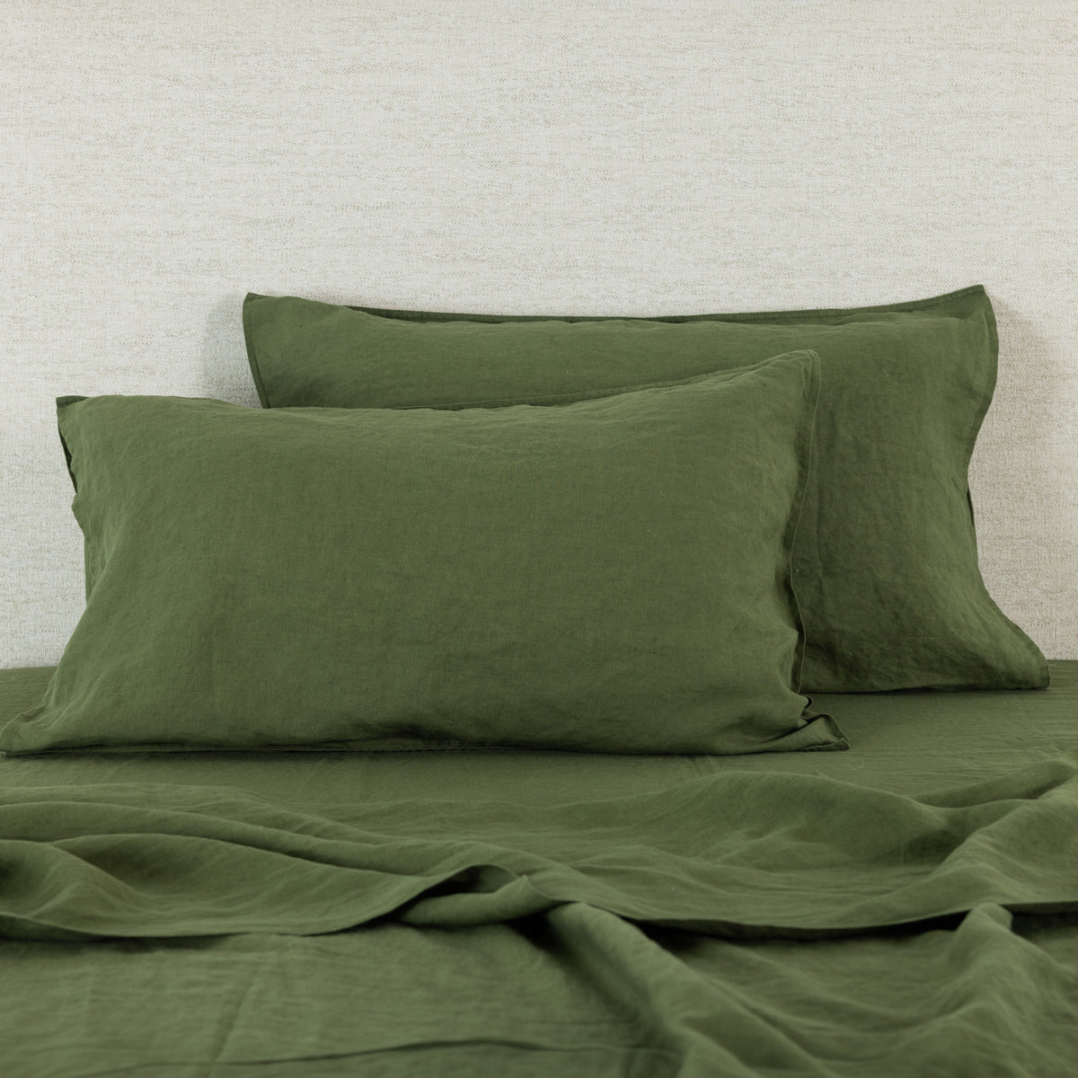 Pine green linen pillowcases are beautifully layered against a neutral textured backdrop, showcasing their natural, relaxed texture and timeless elegance.