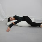 A woman in a black long-sleeve top and black leggings lies on her side on a gray carpeted floor, resting her head on a white crescent-shaped pillow.