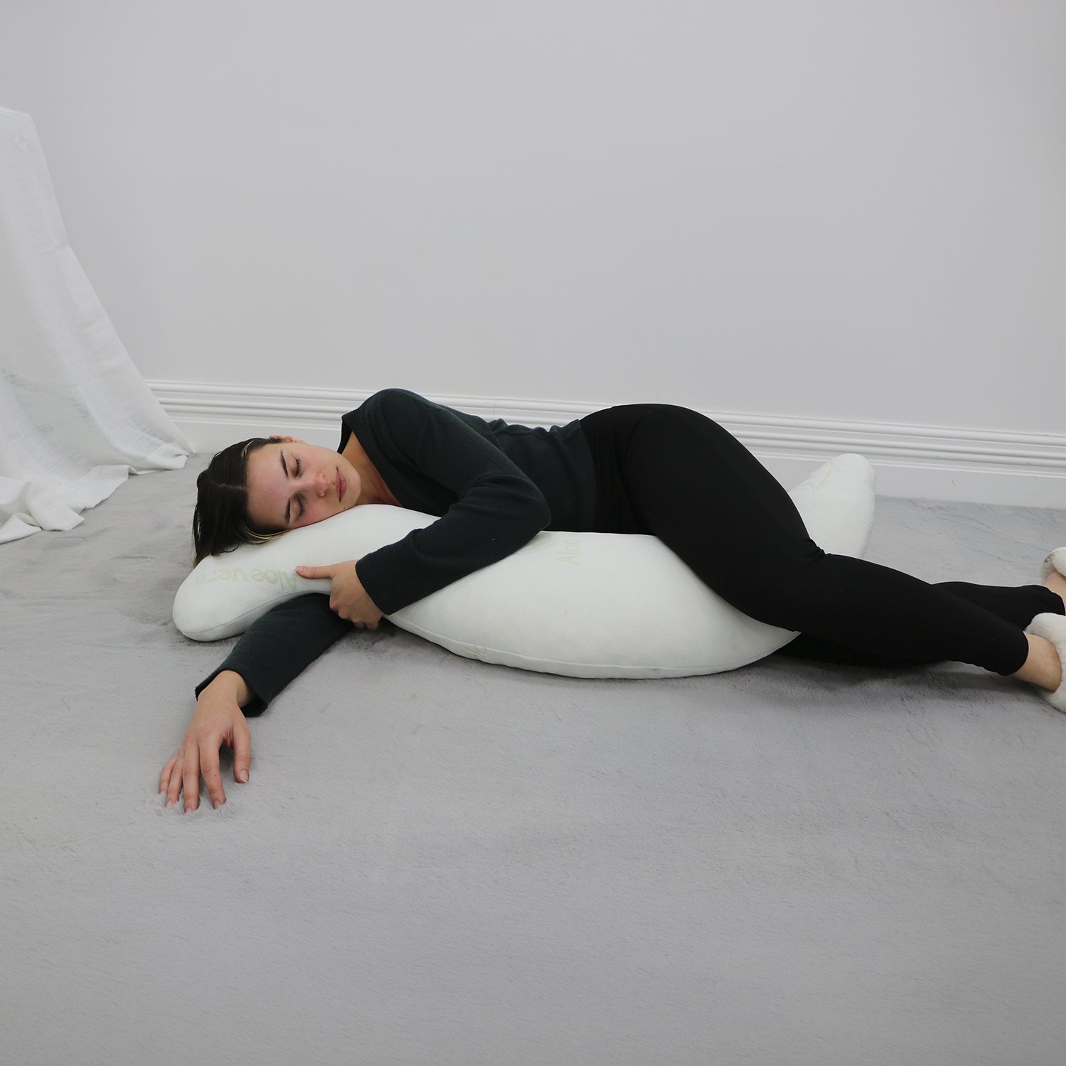 A woman in a black long-sleeve top and black leggings lies on her side on a gray carpeted floor, resting her head on a white crescent-shaped pillow.