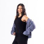 A woman modeling a black sleeveless tank top and black sweatpants, with a dusty blue zip-up hoodie draped off her shoulders against a solid white background.