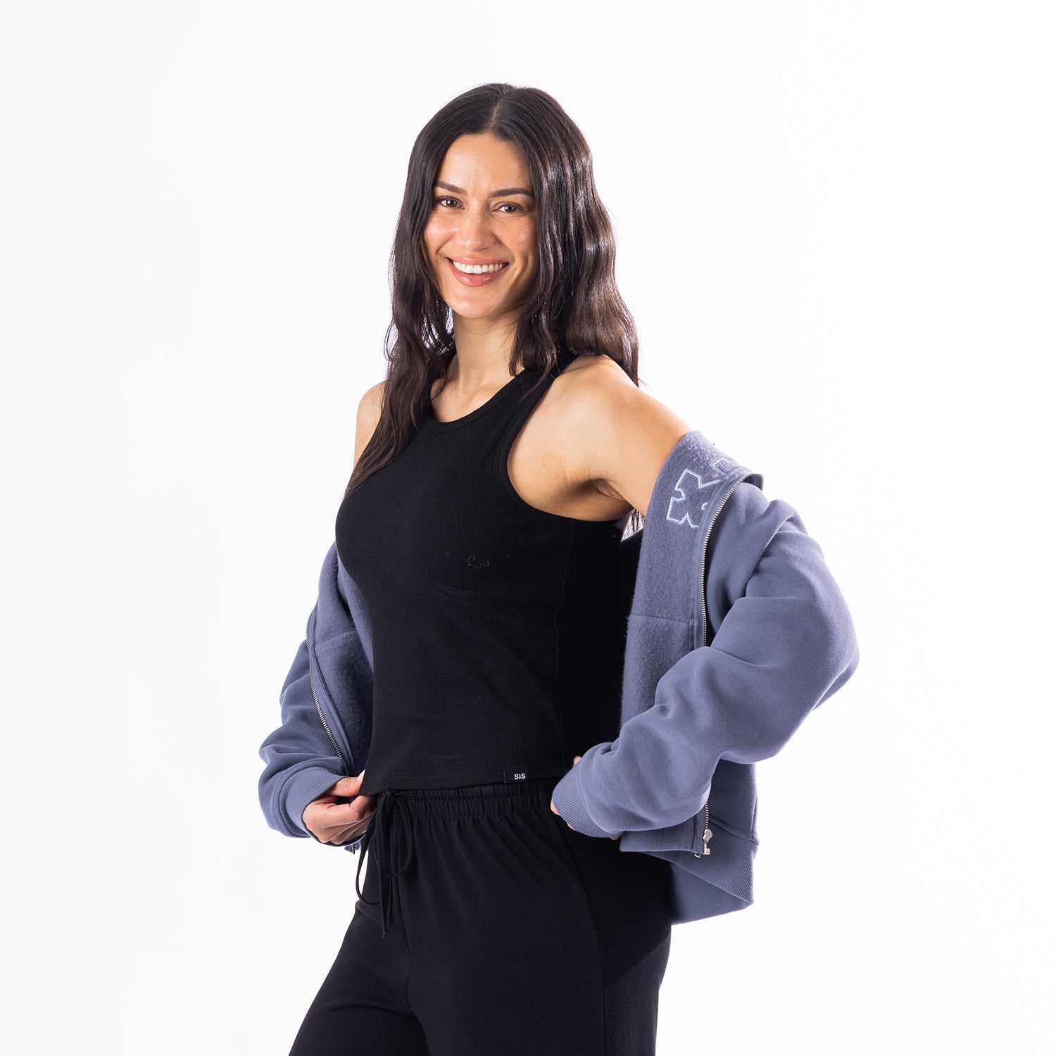 A woman modeling a black sleeveless tank top and black sweatpants, with a dusty blue zip-up hoodie draped off her shoulders against a solid white background.