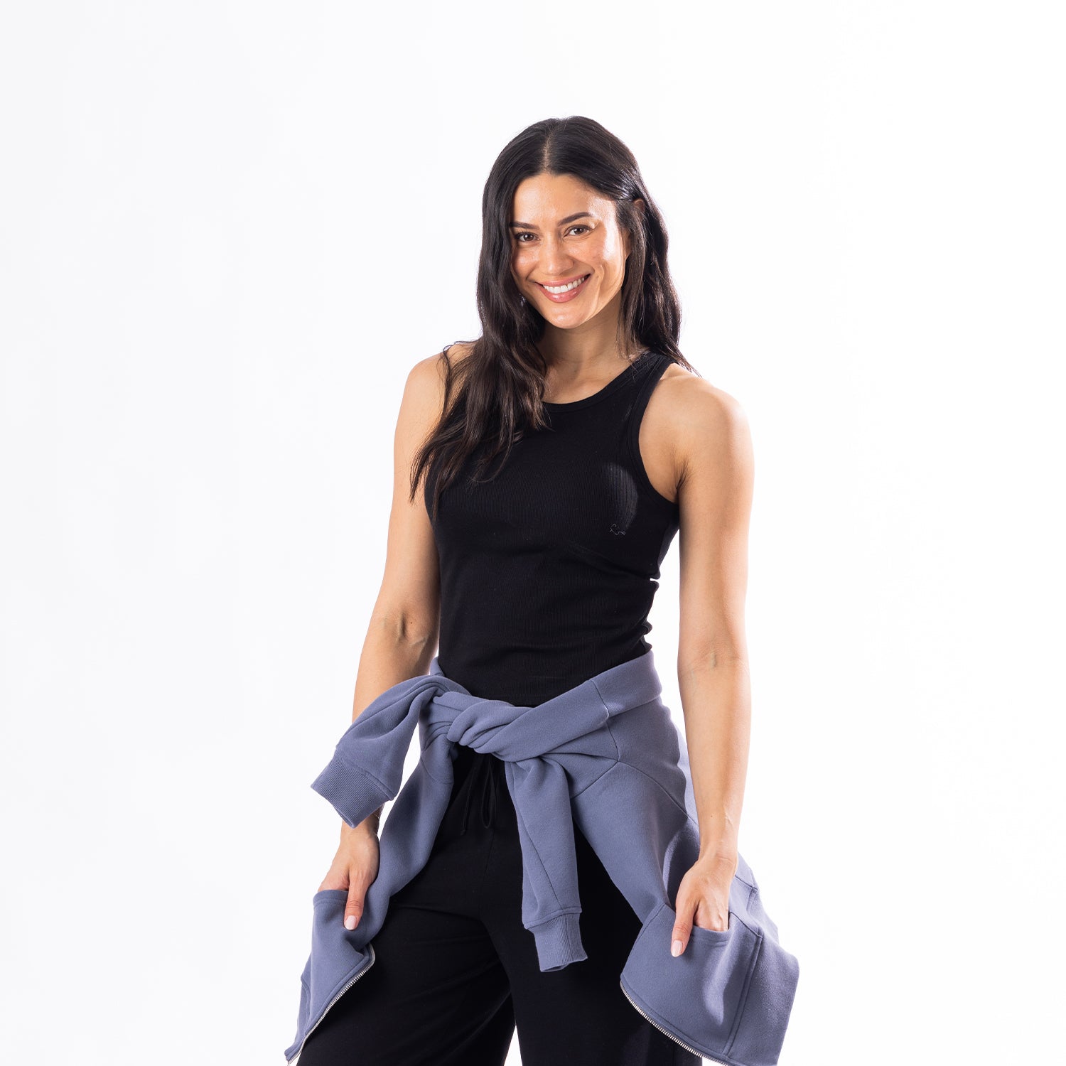 A woman posing in a black athletic tank top and black pants, with a dusty blue sweatshirt tied around her waist, against a solid white background.