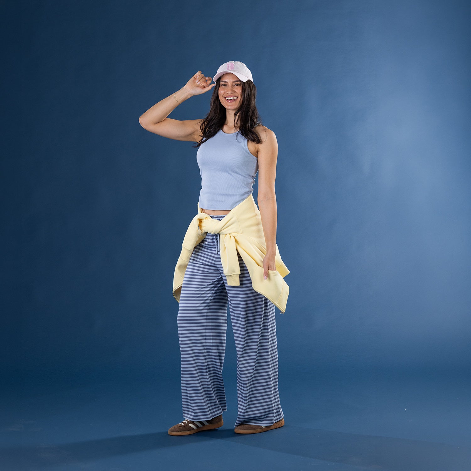 Model wearing the Faded Blue Simple & Sunday Lounge Weekend Ribbed Tank Top with striped lounge pants, a yellow sweatshirt tied around the waist, a white cap and slippers.