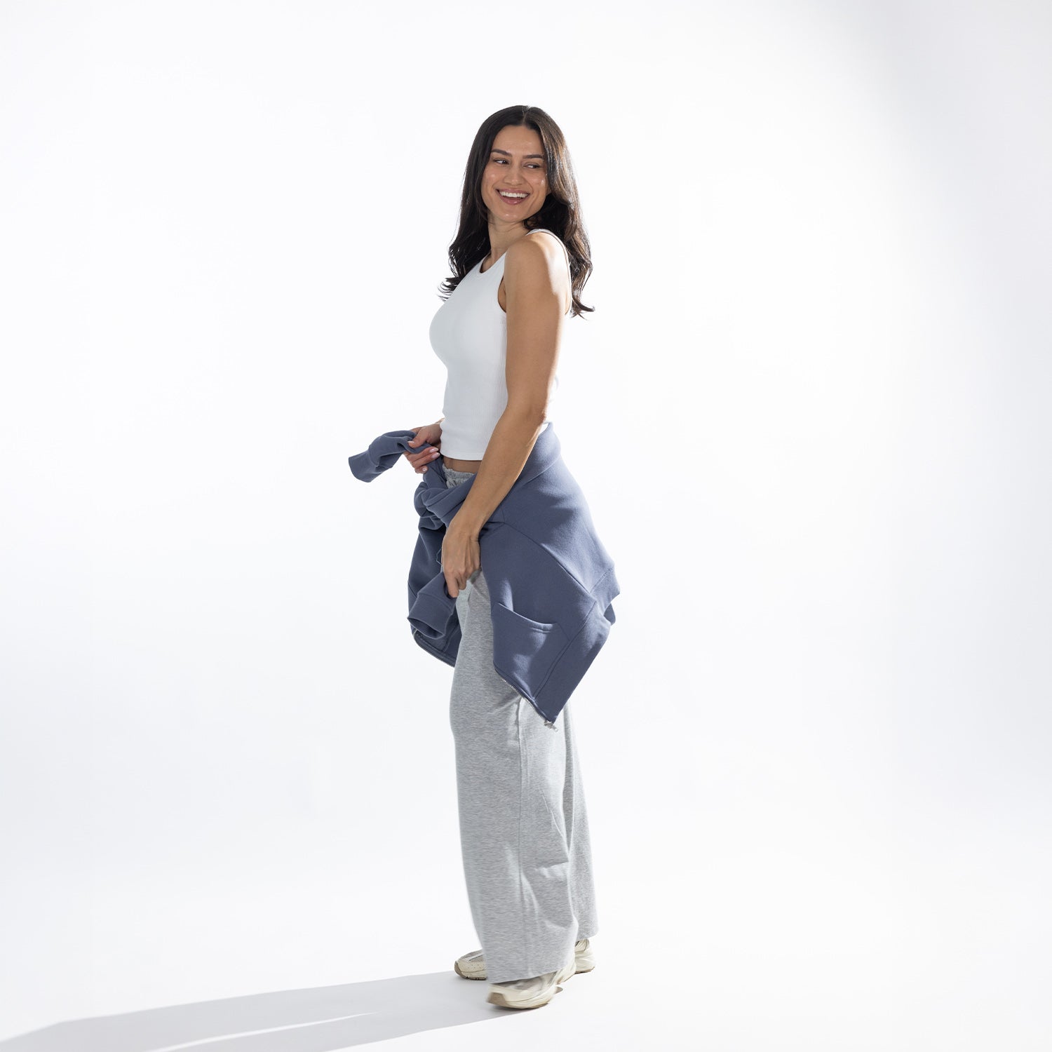 Side profile of the model wearing the White Simple & Sunday Lounge Weekend Ribbed Tank Top with grey wide-leg lounge pants and a blue sweatshirt tied around the waist.