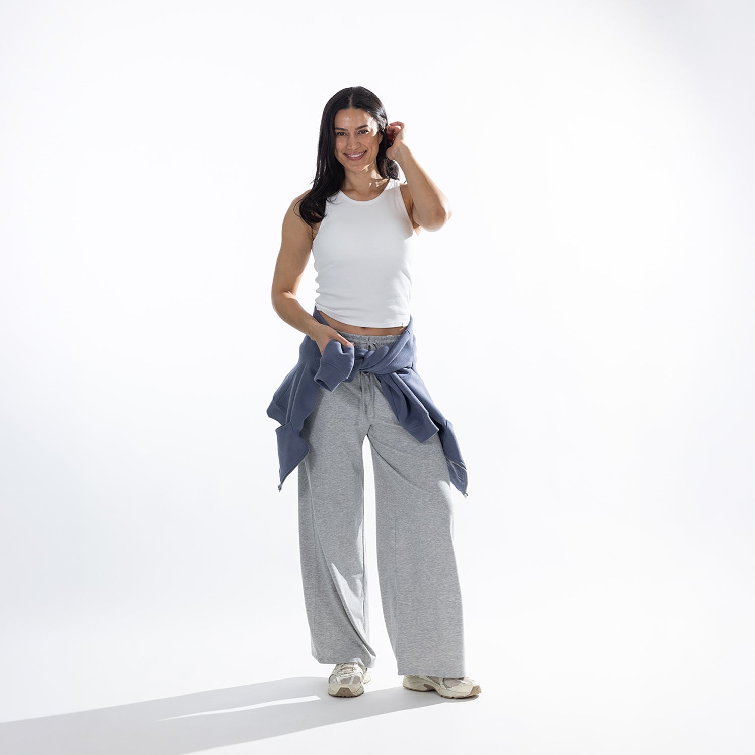 Model wearing the Simple & Sunday Lounge Weekend Ribbed Tank Top in White with grey wide-leg lounge pants, white sneakers and a blue sweatshirt tied around the waist.