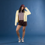 A woman posing in a studio setting against a solid blue backdrop, wearing a light yellow zip-up hoodie, a brown and white striped t-shirt, dark brown shorts, a burgundy baseball cap, and brown high-top sneakers.