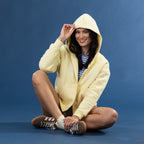 A woman sits cross-legged against a solid blue studio background, wearing a pale yellow zip-up hoodie over a blue and white striped shirt, paired with brown sneakers.