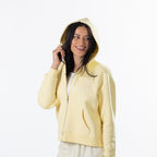 A woman wearing a pale yellow zip-up hoodie with a white graphic print on the chest, posing against a solid white background.