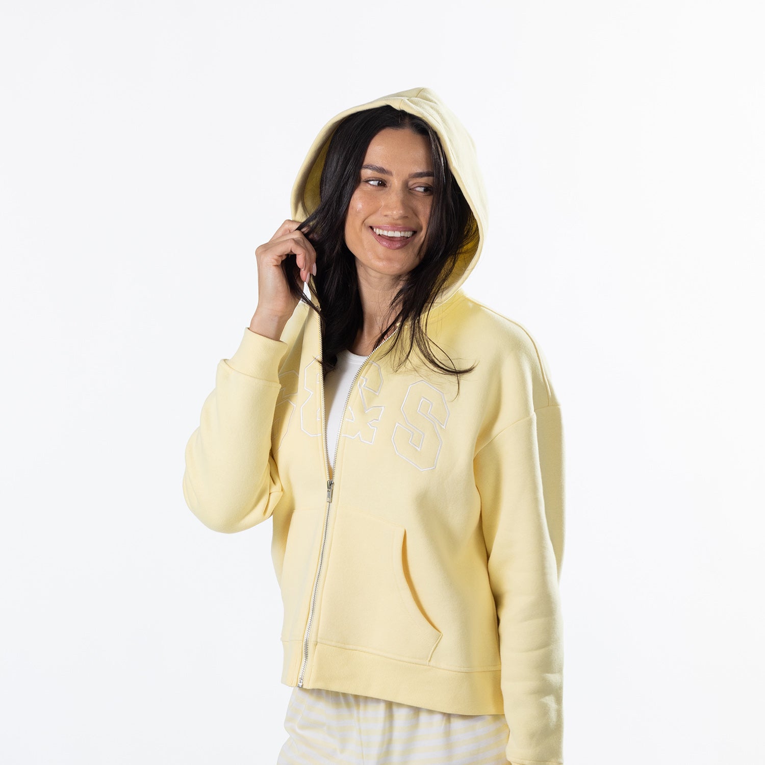 A woman wearing a pale yellow zip-up hoodie with a white graphic print on the chest, posing against a solid white background.