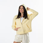 A woman modeling a pale yellow zip-up hoodie with a white t-shirt and striped shorts against a plain white background.