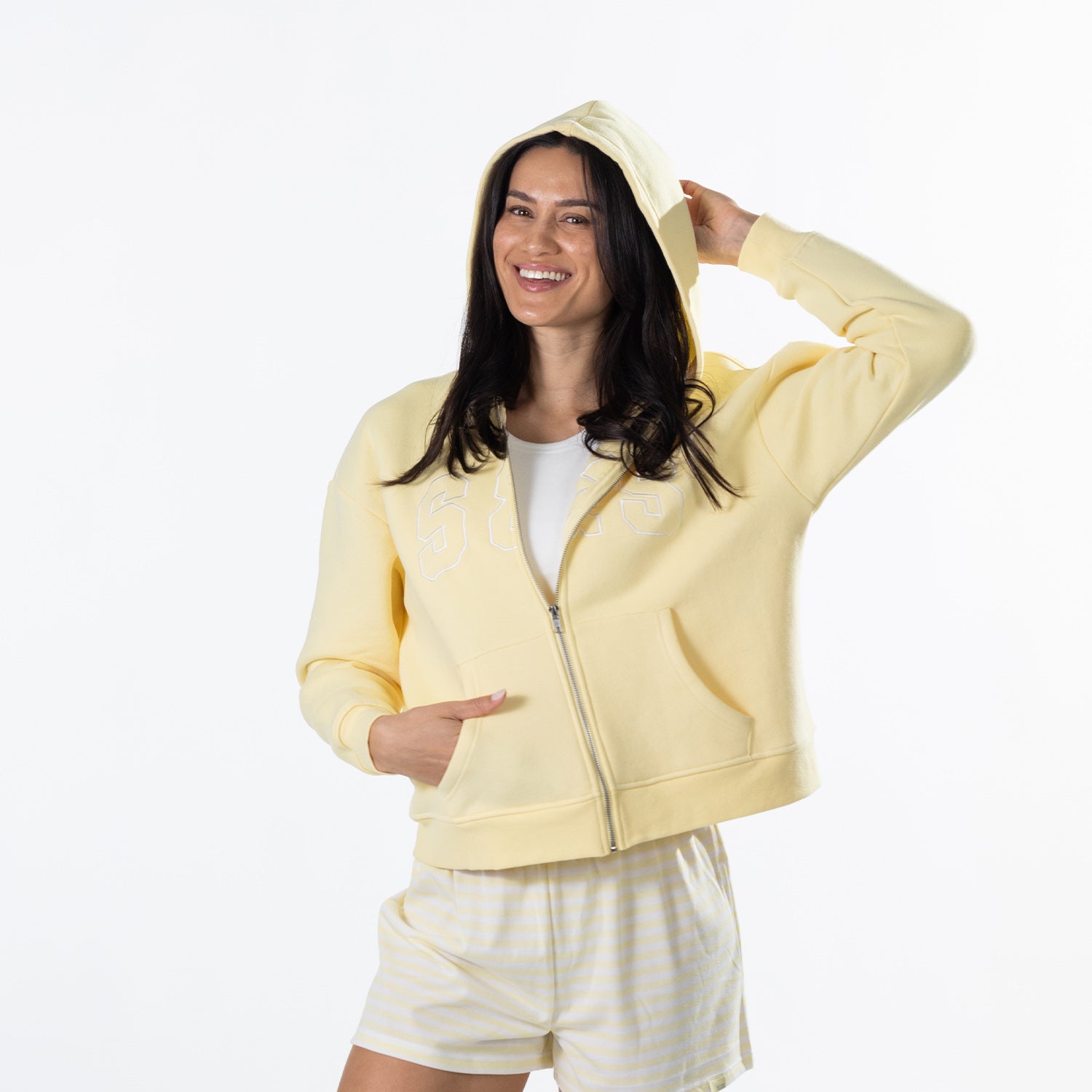 A woman modeling a pale yellow zip-up hoodie with a white t-shirt and striped shorts against a plain white background.