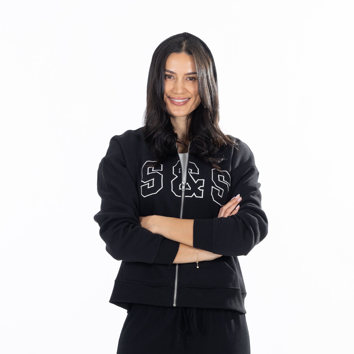 A woman with long dark hair stands against a solid white background, wearing a black zip-up hoodie with 'S&S' printed in white  letters across the chest, paired with black sweatpants.