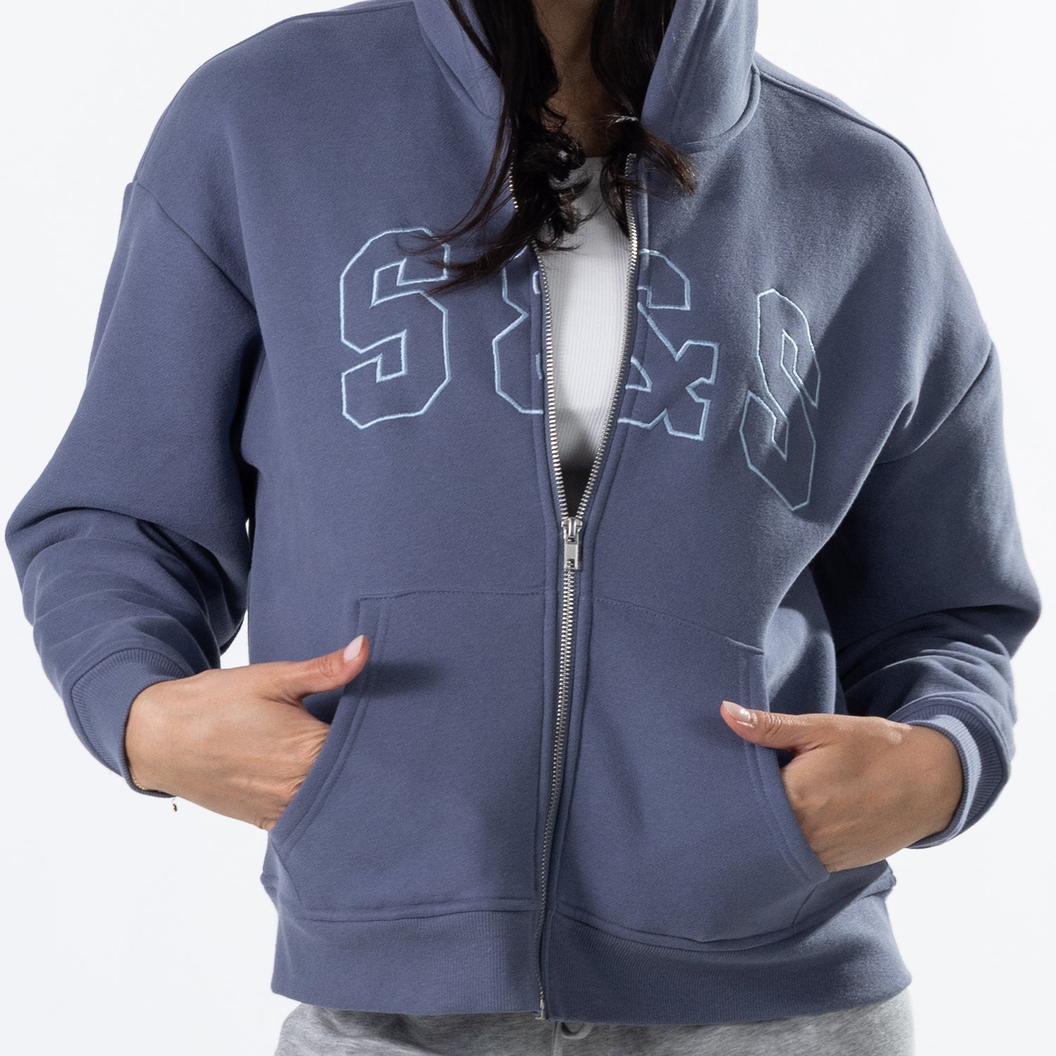 A person wearing a slate blue zip-up hoodie with white outlined 'S&S' lettering across the chest, shown from the neck down against a light gray background.