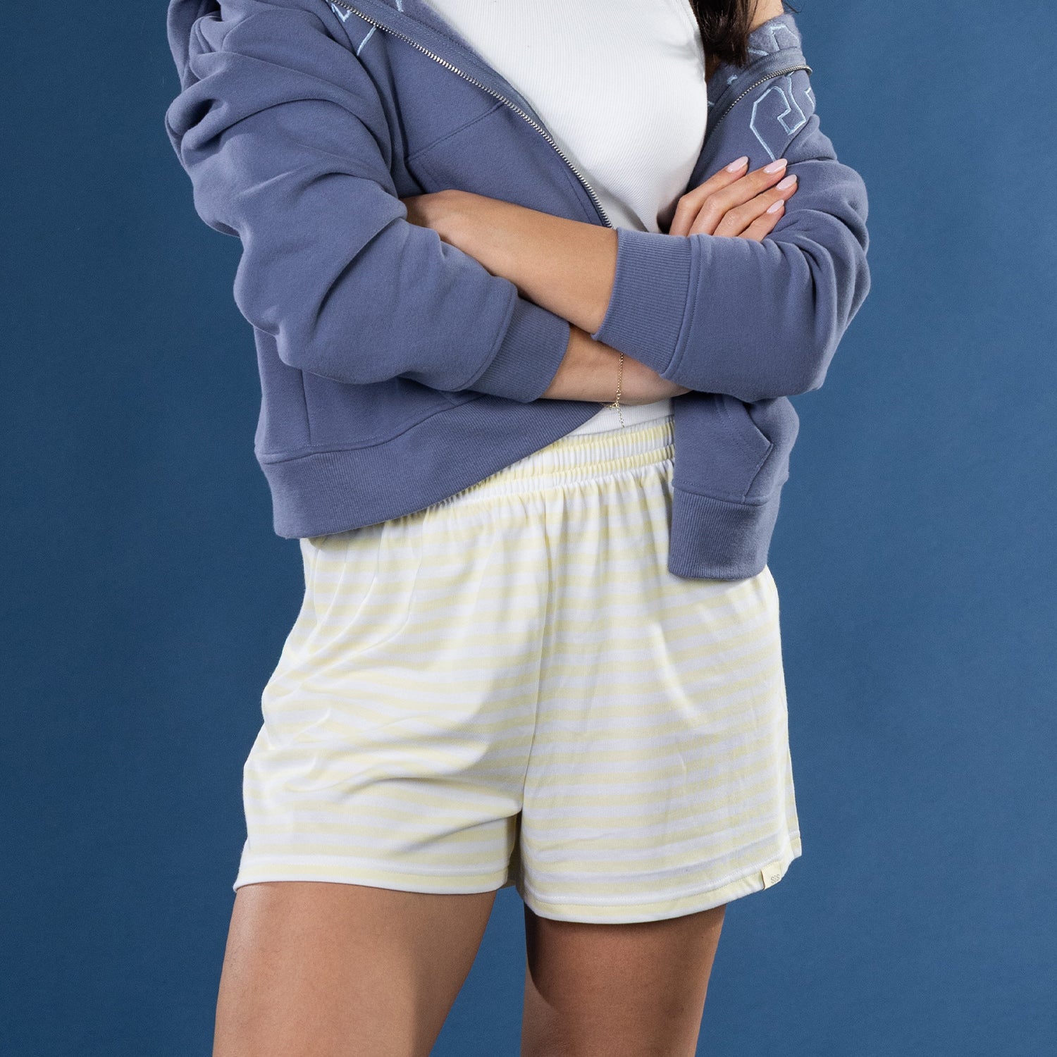 Close-up of the yellow striped lounge shorts on the model with a white ribbed tank top and blue zip hoodie.