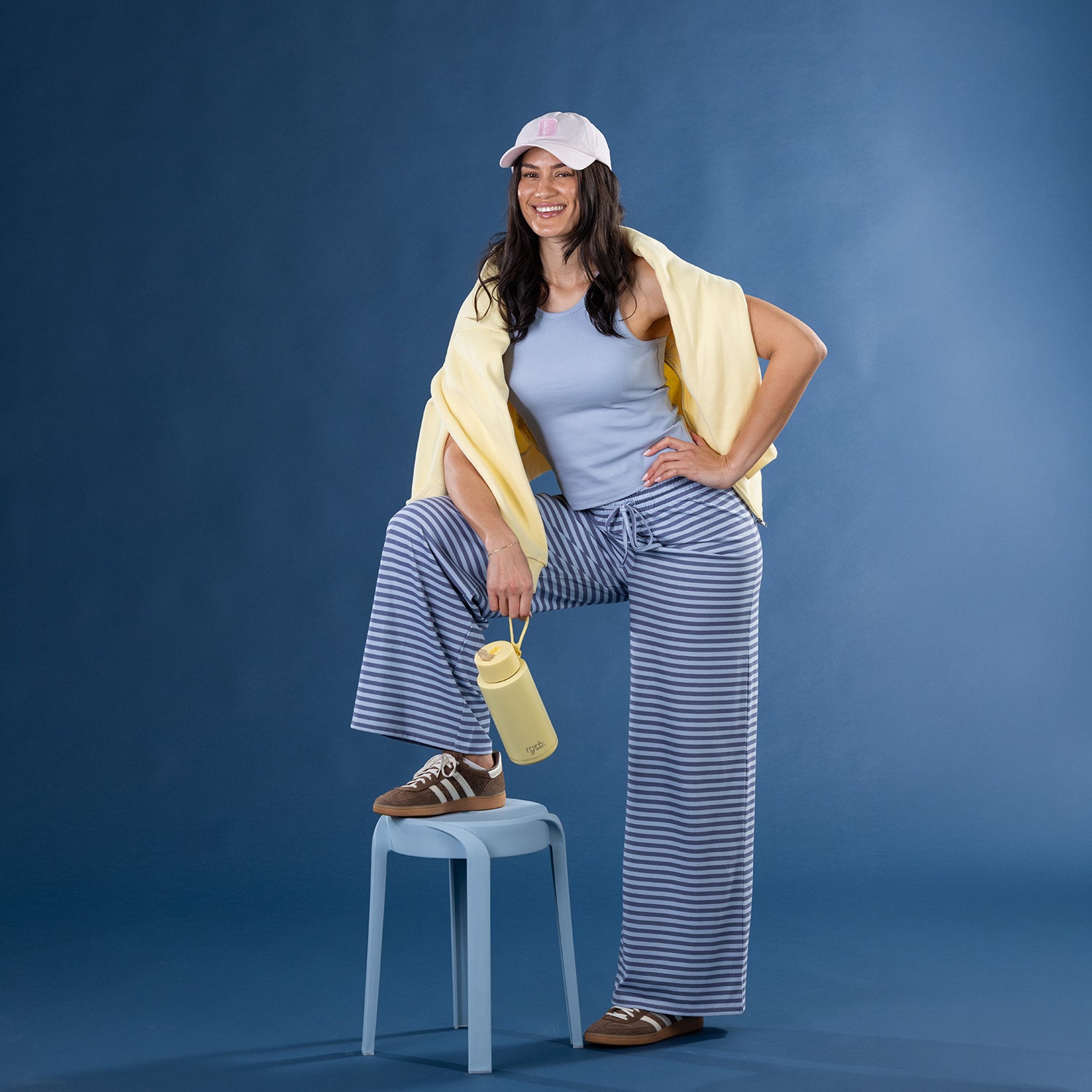 Model wearing faded blue striped lounge pants with a blue ribbed tank top and yellow zip hoodie draped over the shoulders.