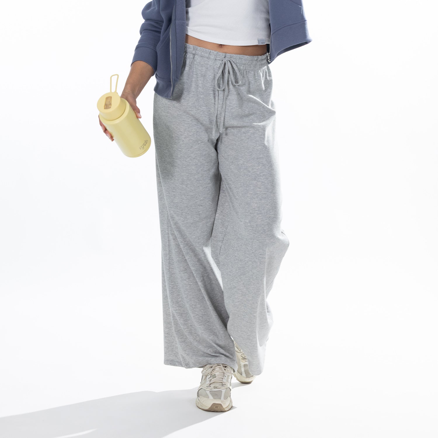 A person wearing loose-fit  grey drawstring sweatpants, a blue zip-up cardigan, a white crop top, and beige sneakers, holding a pale yellow tumbler against a clean white studio background.