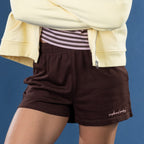 A person wearing dark brown lounge shorts with a white script logo on the lower left hem, styled with a striped waistband and a pale yellow zip-up hoodie against a blue background.