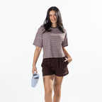 A woman modeling a brown and beige horizontally striped short-sleeve crop top and dark brown athletic shorts, holding a light blue water bottle against a plain white background.