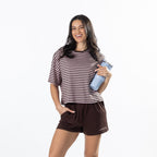 A woman modeling a brown and dark pink striped t-shirt and dark brown shorts, holding a light blue water bottle against a solid white background.