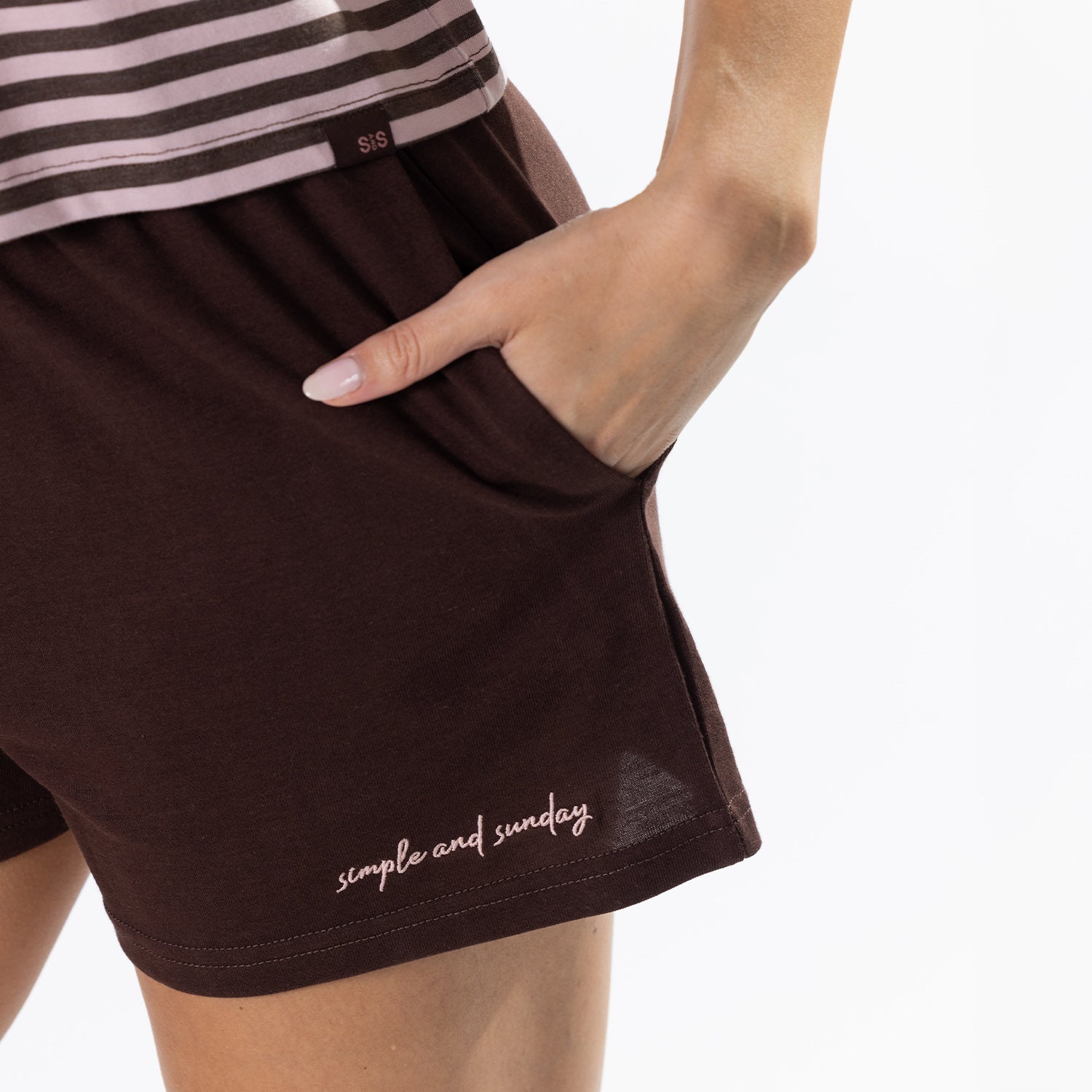 Close-up of a person wearing dark brown shorts with a side pocket, hand in pocket, and 'simple and sunday.' printed near the hem on a white background.