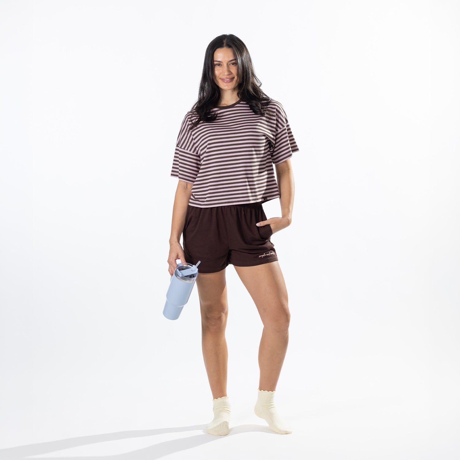 A woman modeling a brown and white striped short-sleeve t-shirt and dark brown athletic shorts, holding a light blue water bottle against a clean white studio background.