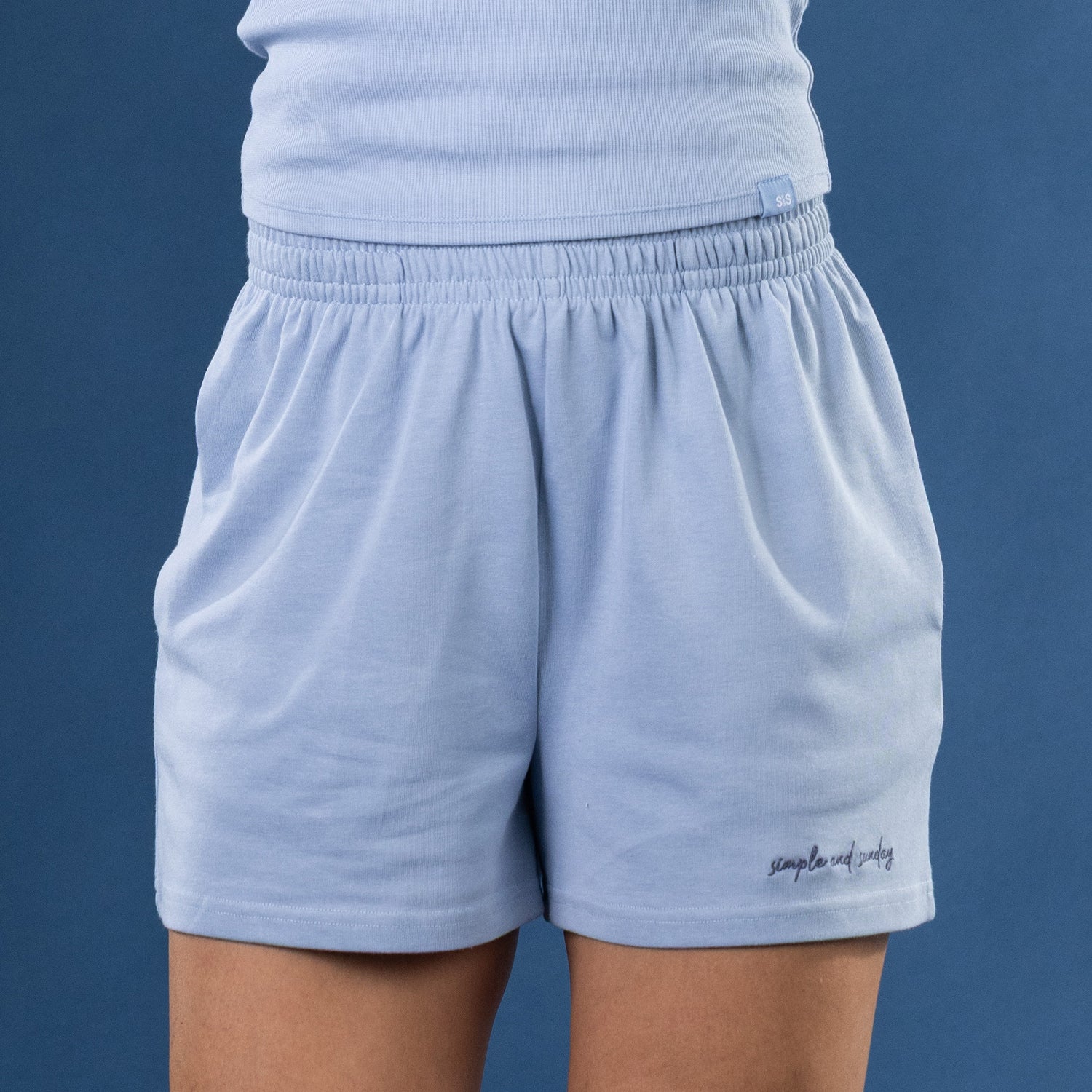 Light blue athletic shorts with an elastic waistband, shown on a model against a solid blue background. The brand name 'simple and Sunday' is embroidered in black script on the lower left leg.