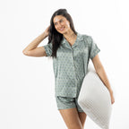 Woman modelling the sage printed satin pyjama set with a short-sleeve button-up top, revere collar and matching shorts while holding a pillow.