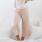 A person wearing light pink and white floral cropped pajama pants, standing on a white bed with pillows.