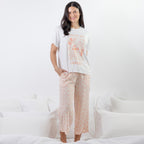 A woman modeling a two-piece pajama set consisting of a white graphic t-shirt and light pink floral cropped pants, standing on a white bed.