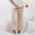 A person wearing light peach and white floral cropped pants with a white t-shirt, standing on a white bed with textured pillows.