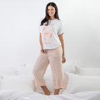 A woman modeling a two-piece pajama set consisting of a white graphic t-shirt and light pink floral cropped pants, standing on a bed with white bedding.