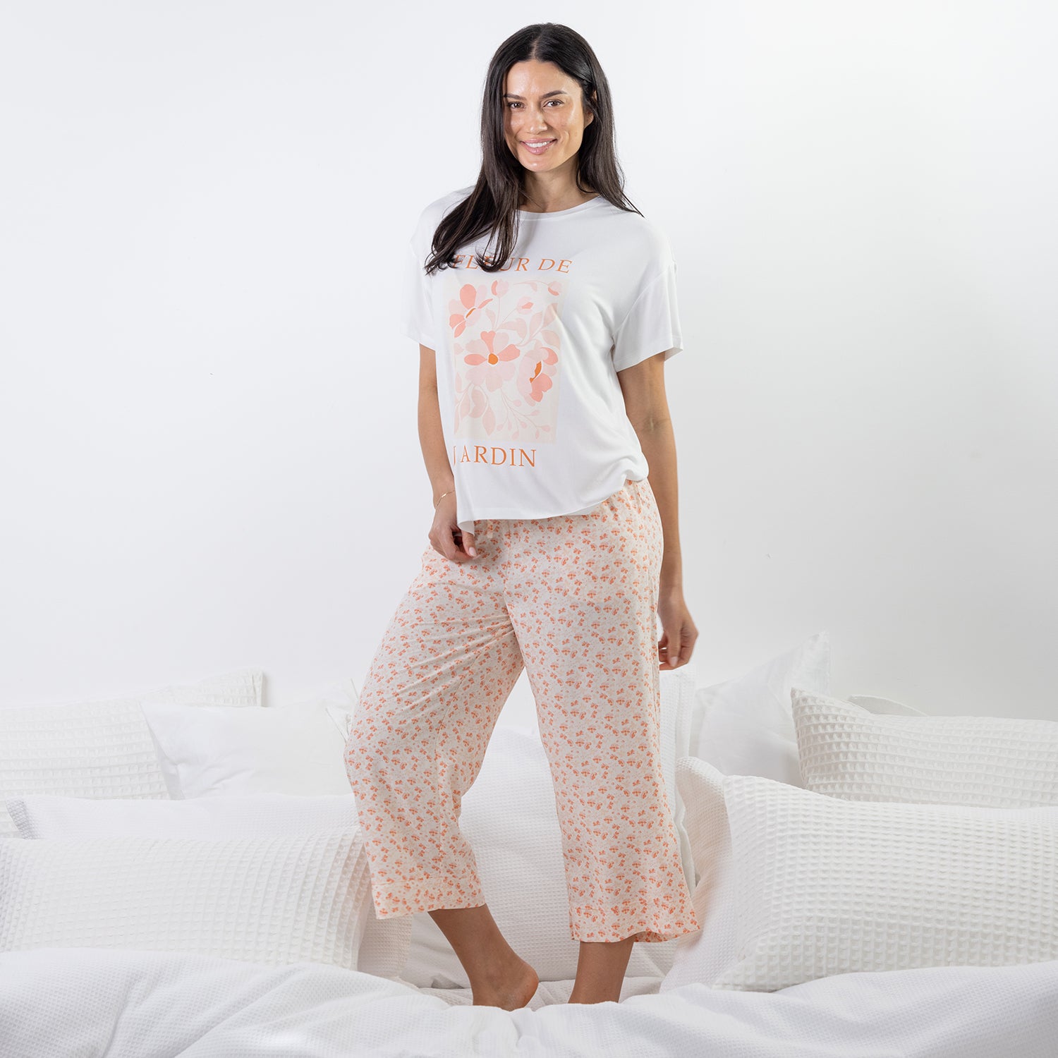 A woman modeling a two-piece pajama set consisting of a white graphic t-shirt and light pink floral cropped pants, standing on a bed with white bedding.