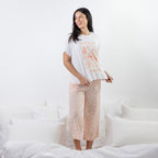 A woman modeling a two-piece pajama set consisting of a white short-sleeve t-shirt with a pink floral graphic and matching light pink patterned cropped pants, standing on a bed with white bedding.
