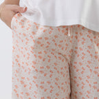 Close-up of a person wearing light beige shorts with a small orange bow pattern, paired with a white top. A hand is visible pulling the hem of the top to show the shorts' fit.