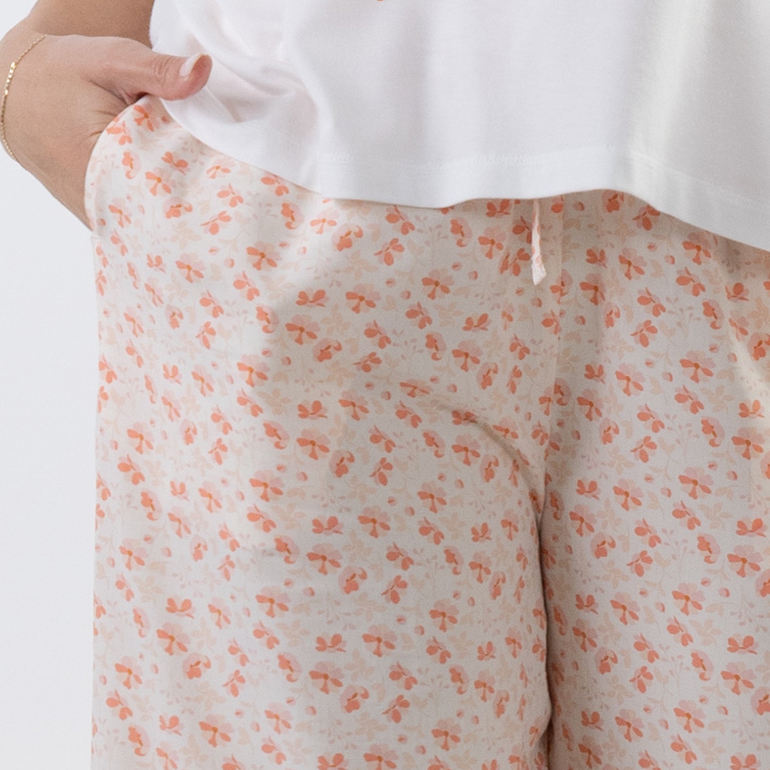 Close-up of a person wearing light beige shorts with a small orange bow pattern, paired with a white top. A hand is visible pulling the hem of the top to show the shorts' fit.
