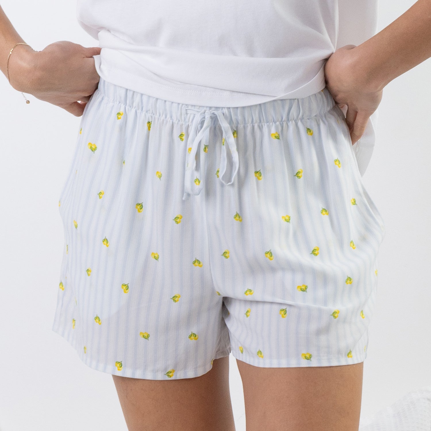White drawstring shorts with thin vertical blue stripes and small yellow lemon motifs, shown on a model wearing a white top against a light gray background.