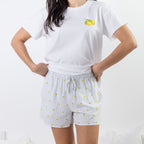 A woman wearing a white short-sleeve t-shirt with a small yellow lemon graphic on the left chest and white pinstriped shorts with a yellow lemon print, standing with hands on hips against a white background.
