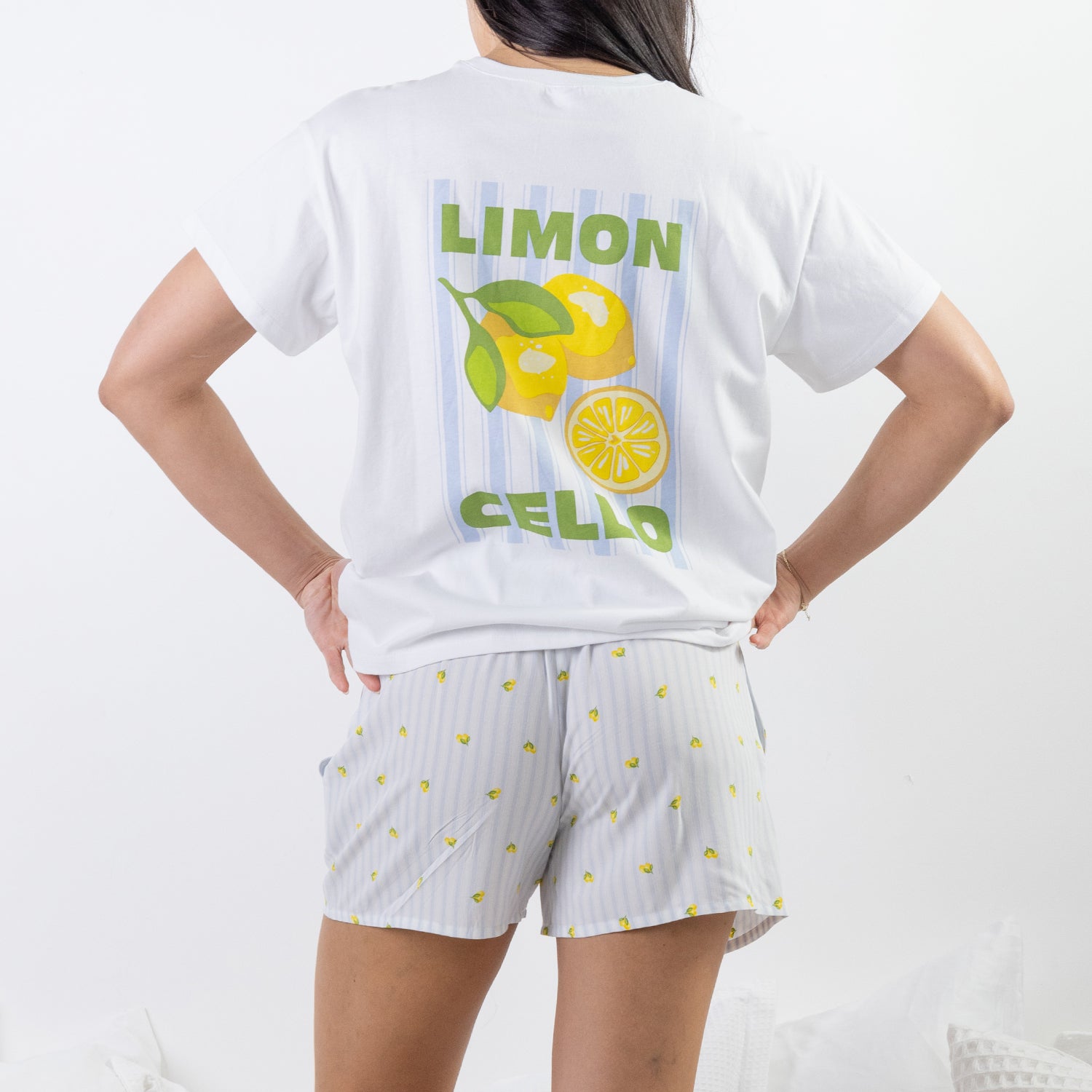 A woman shown from the back wearing a white short-sleeve T-shirt and matching white shorts with small yellow lemon prints. The shirt has a large back graphic with green text reading 'LIMON CELLO' and a yellow lemon illustration on a light blue striped background.