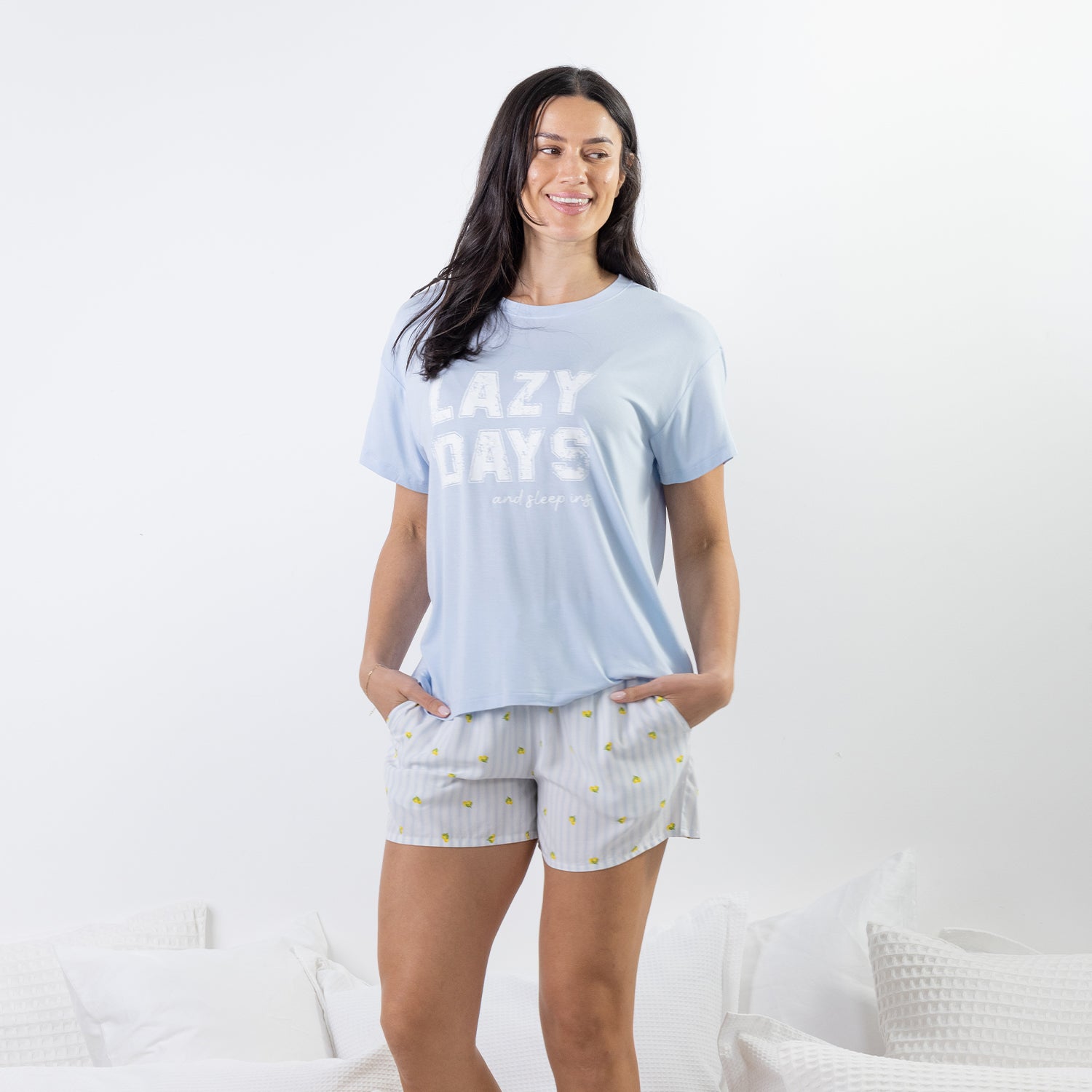 A woman modeling a light blue t-shirt with 'LAZY DAYS' printed in white, paired with white and blue striped shorts with yellow sun prints, standing in a brightly lit bedroom setting.