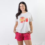 A woman modeling a white short-sleeve t-shirt with a colorful graphic of pancakes, strawberries, and a red drink, paired with pink and white striped shorts, against a plain white background.