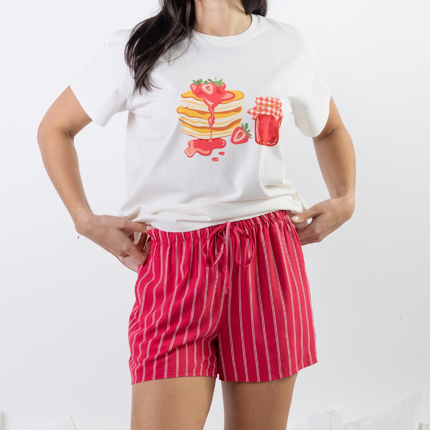 A woman wearing a white graphic t-shirt with a strawberry pancakes print and red-and-white vertically striped shorts, standing against a plain white background.