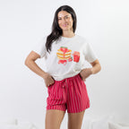 A woman modeling a white graphic t-shirt with a breakfast-themed print (pancakes, strawberries, coffee cup) and red-and-white vertically striped shorts, set against a clean white studio background.