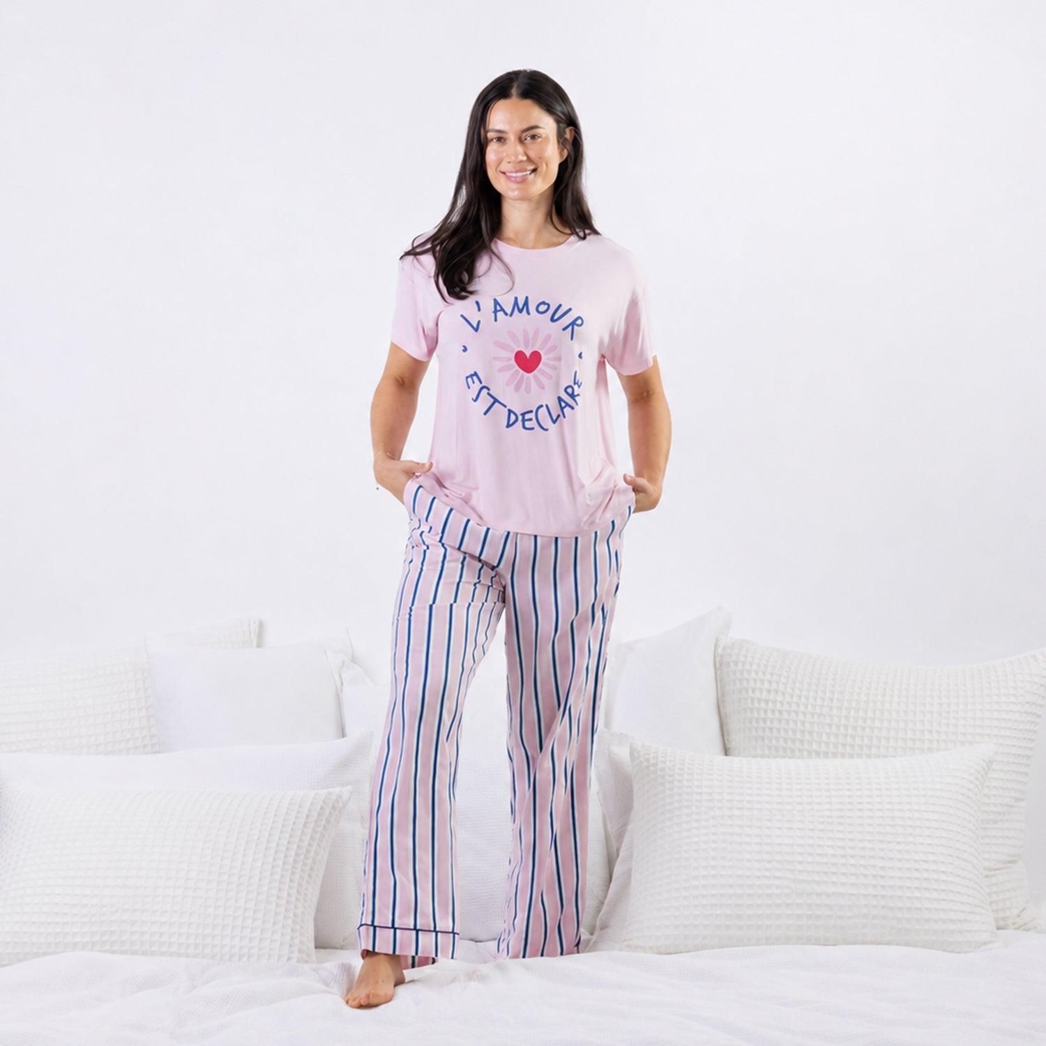 A woman stands on a bed wearing a light pink pajama set with a blue graphic text design on the top and blue and pink vertical stripes on the pants.