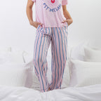 A person wearing a pink graphic T-shirt and pink pants with vertical blue and white stripes, standing on a white bed with pillows.